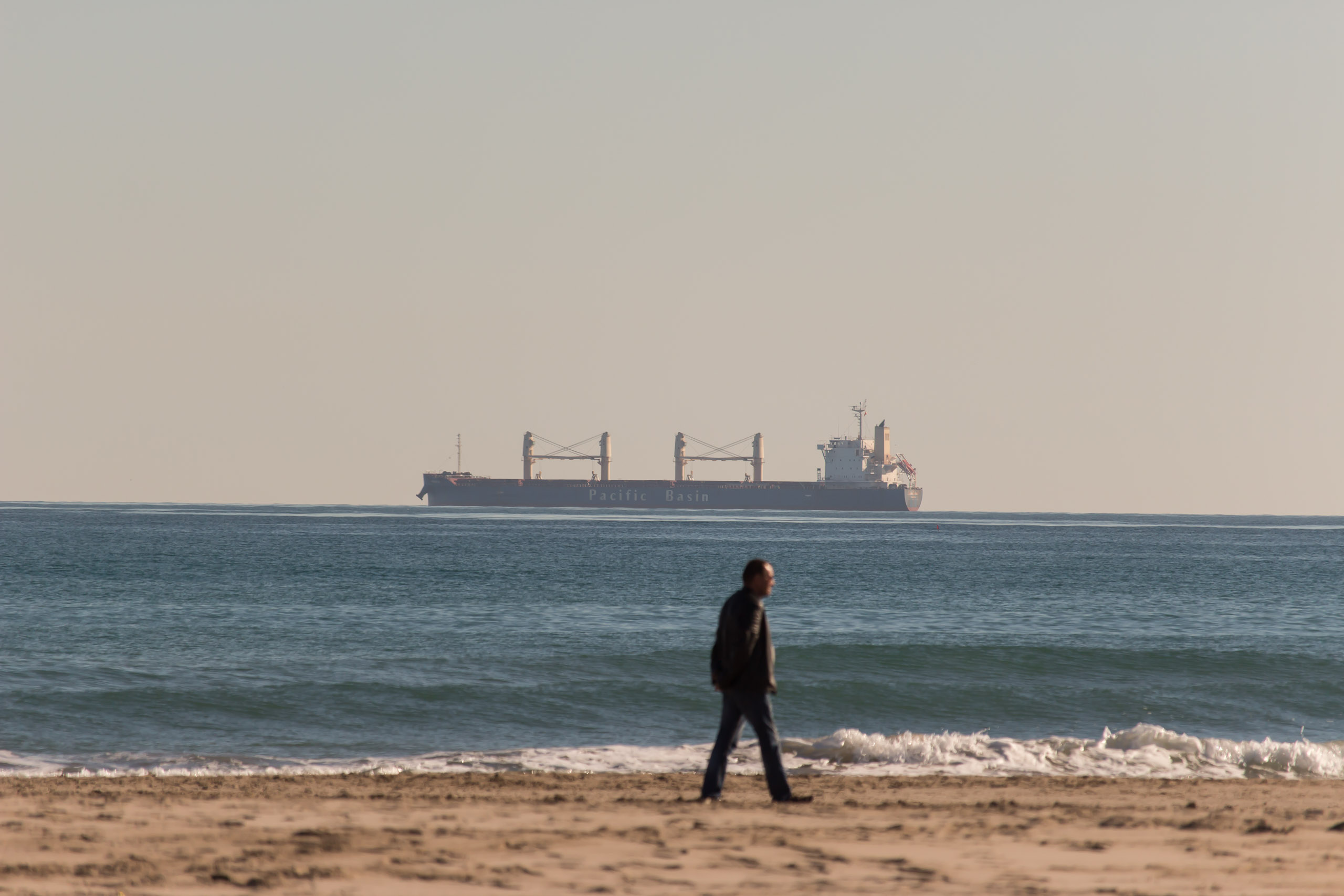Ein Mann spaziert am Strand, im Hintergrund ist ein Tanker 